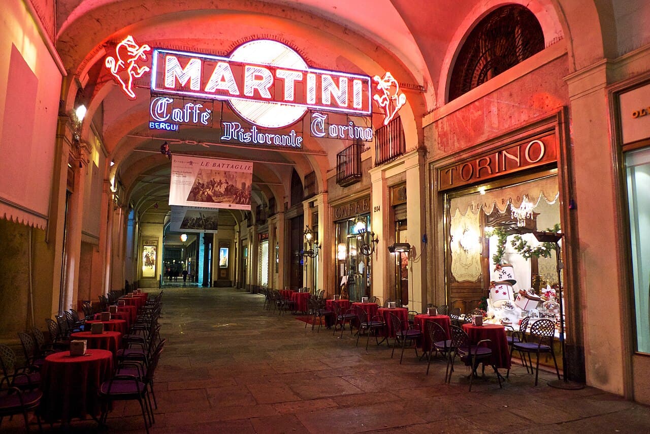 A historic arcade at night: café tables set out under the vaults beside the neon sign of a Martini caffè