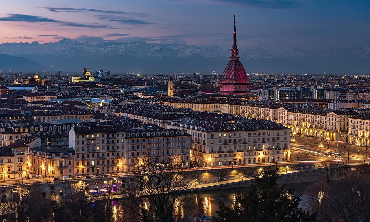 Turin at dusk seen from the hill — the Mole Antonelliana lit red above lamplit boulevards, the Po in the foreground and the snow-covered Alps along the horizon