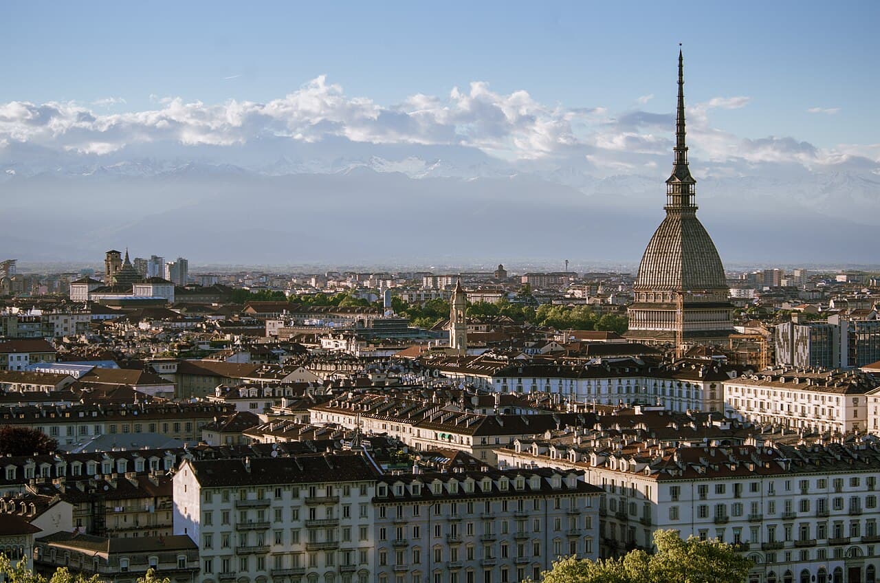 Turin's rooftops in afternoon light, the Mole Antonelliana rising on the right and the snow-capped Alps behind the city
